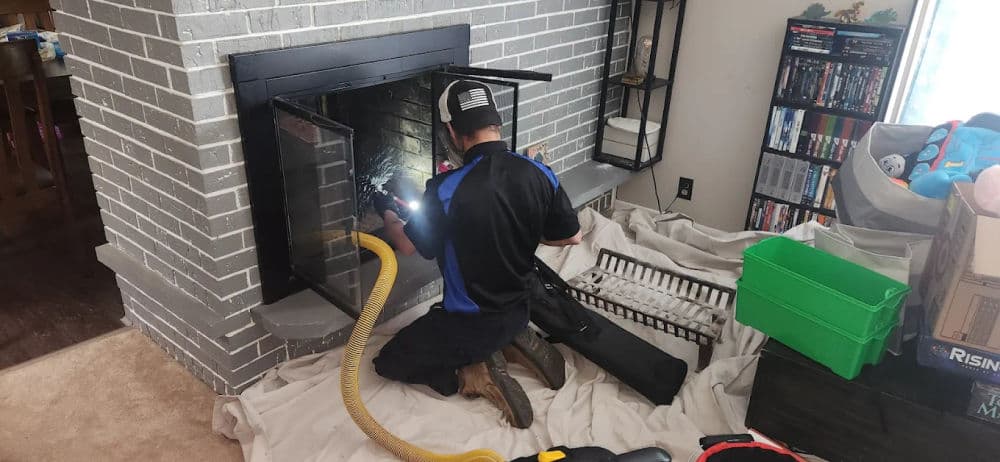 Technician cleaning a fireplace with tools, wearing a cap, in a home setting.